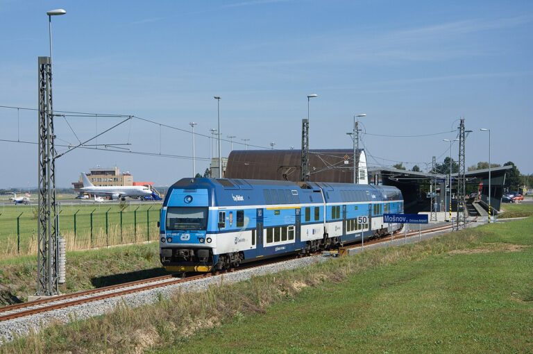 Modrá elektrická jednotka odjíždí ze železniční stanice u letiště Ostrava v Mošnově, v pozadí budova terminálu